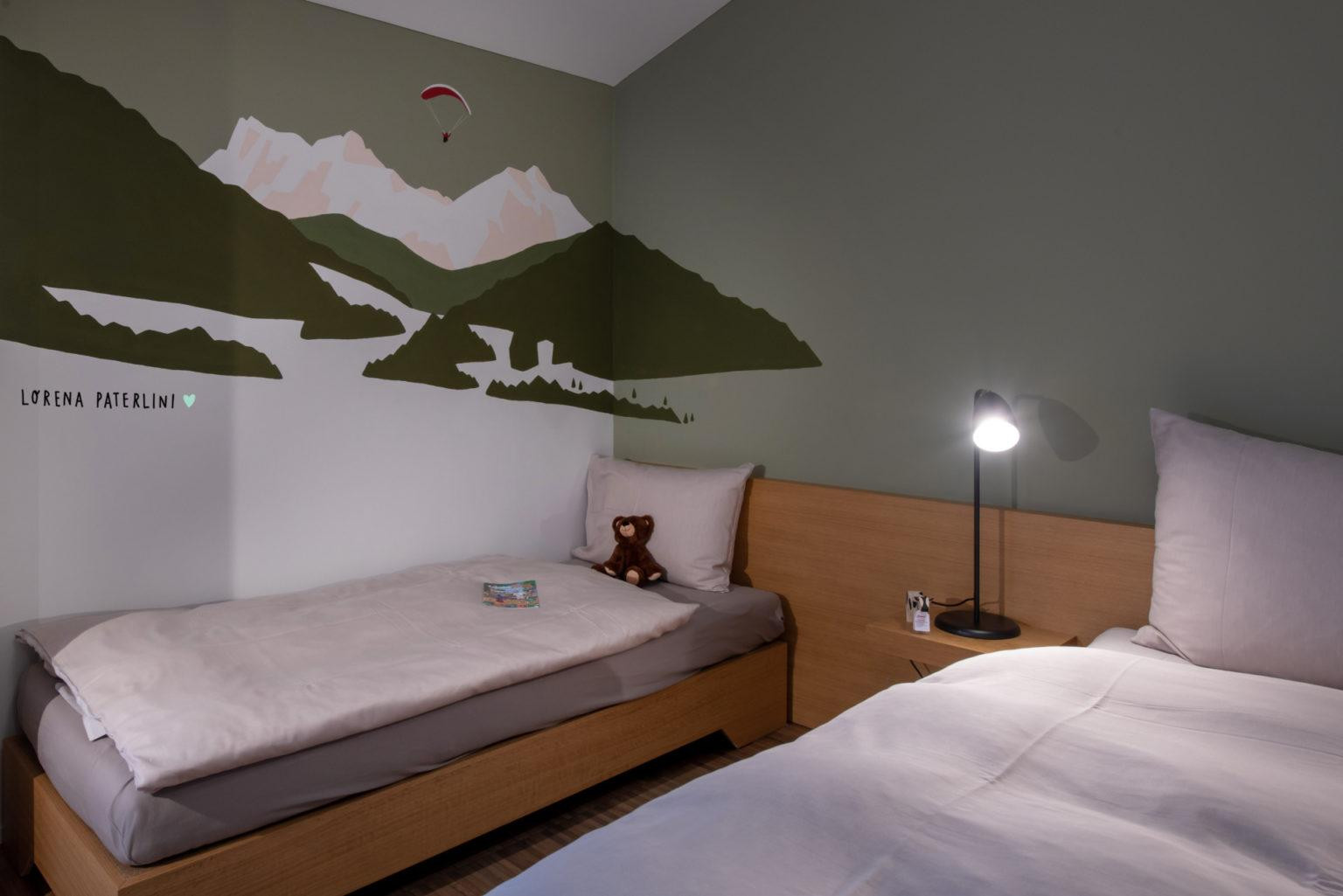 Budget Zimmer mit gemütlichem Doppelbett und Einzelbett mit einem Teddybären und Wandillustration mit alpinen Skimotiven.